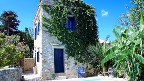 A stone building with a blue door and windows, partially covered in green vines, adjacent to a swimming pool. - Villa Sofas (Platanias)