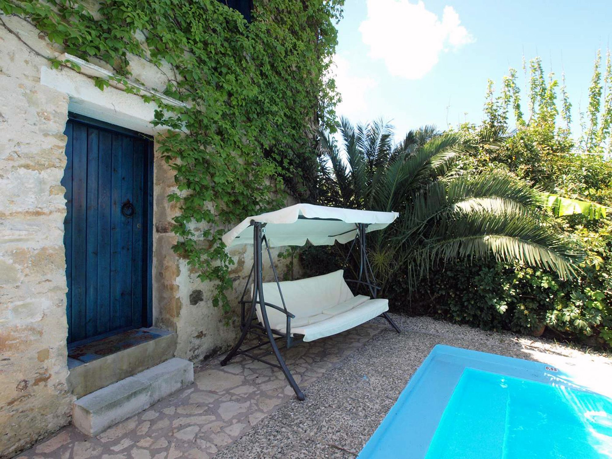 A stone building with a blue door, a white swing chair, and a pool.