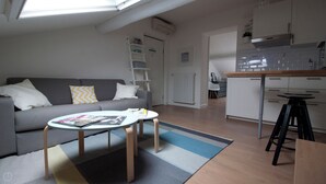 Uma sala de estar moderna com um sofá cinza, uma mesa de centro e uma área de cozinha com armários brancos e bancada de madeira.