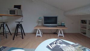 Quarto pequeno e moderno, com uma escrivaninha de madeira, televisão, dois bancos altos, uma estante e uma revista no chão.