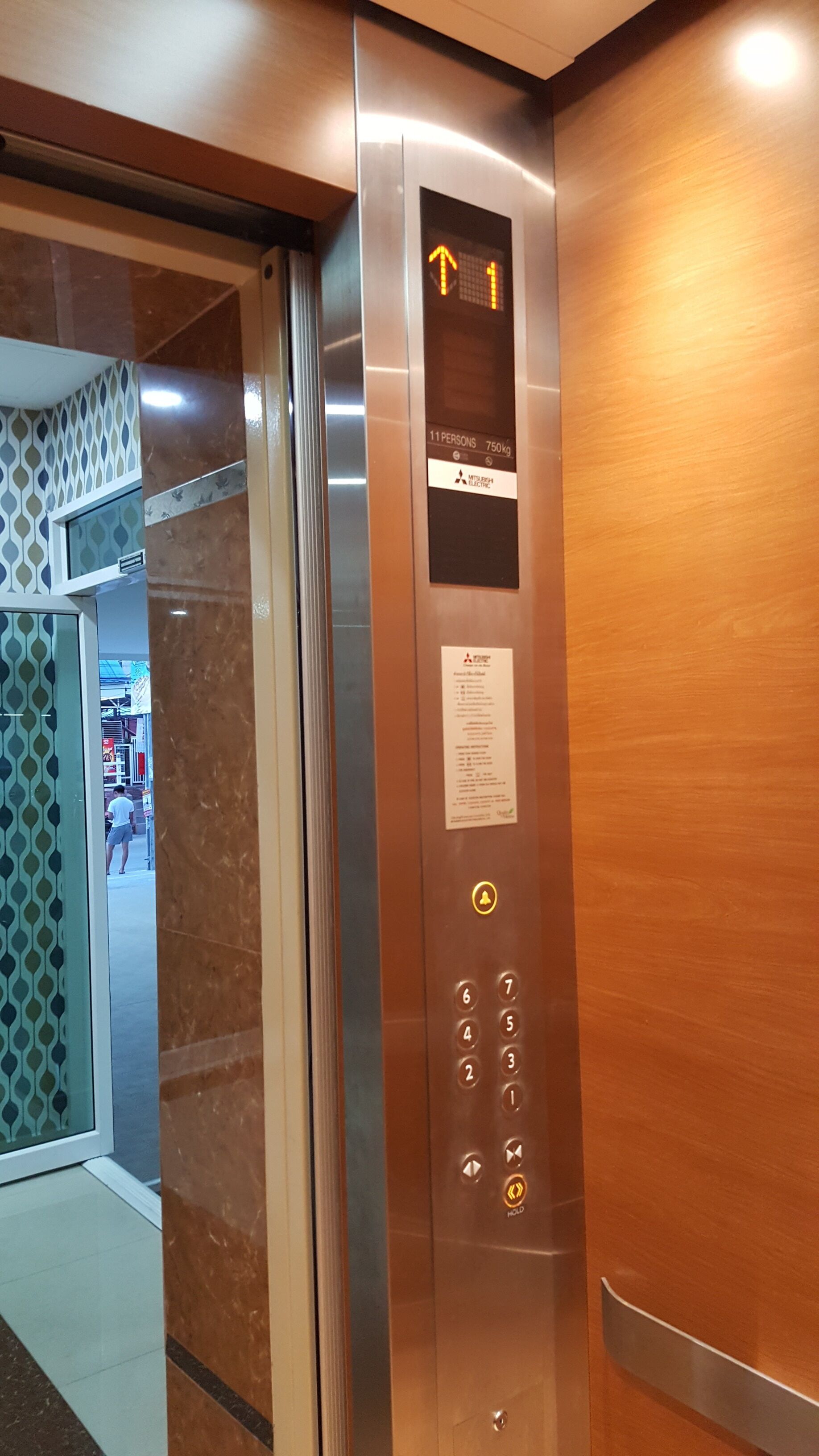 Interior de un ascensor con pantalla digital que muestra el número de piso, un cartel con el límite de capacidad y una lista de servicios.