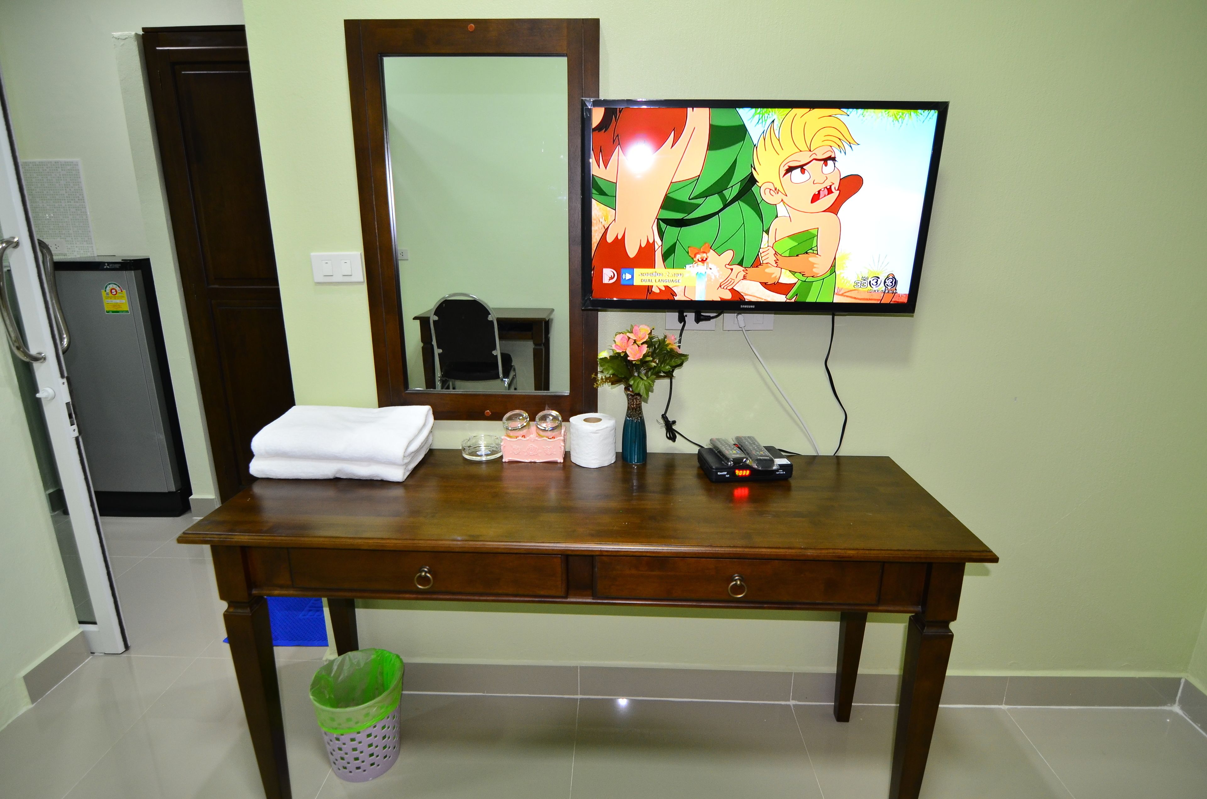 Una habitación de hotel con un escritorio de madera, un televisor montado en la pared, un espejo sobre el escritorio y un cesto de basura debajo.