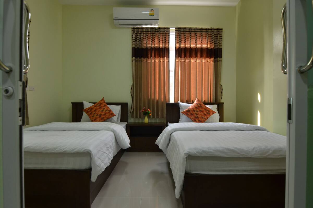 A hotel room with two beds, a nightstand, a window with curtains, and an air conditioning unit.