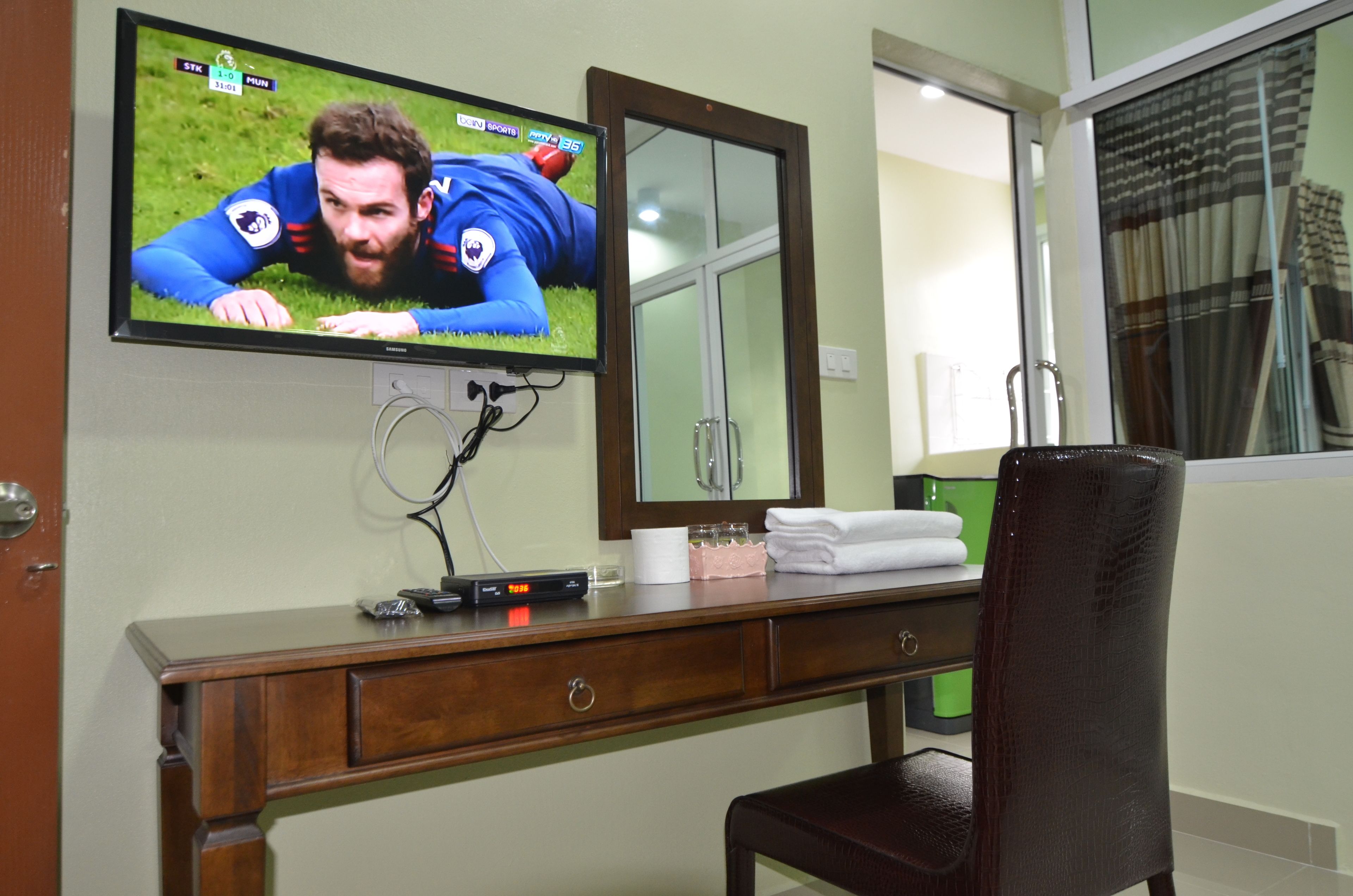 A hotel room with a TV mounted on the wall displaying a sports event, a wooden desk with a mirror, a chair, and a window with curtains.