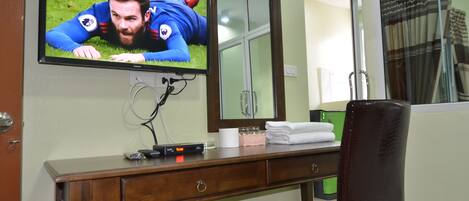 A hotel room with a TV mounted on the wall displaying a sports event, a wooden desk with a mirror, a chair, and a window with curtains.