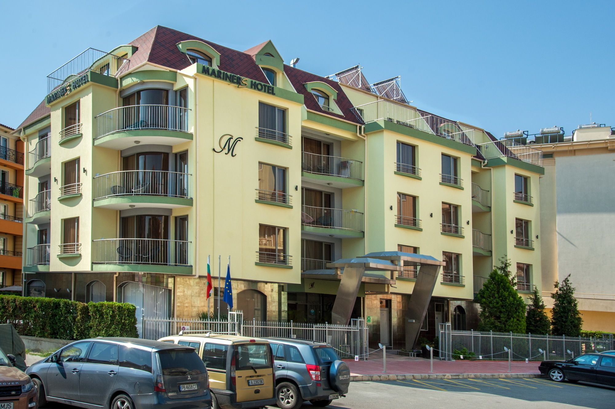 A multi-story hotel with balconies, a central entrance, and a parking area in front.