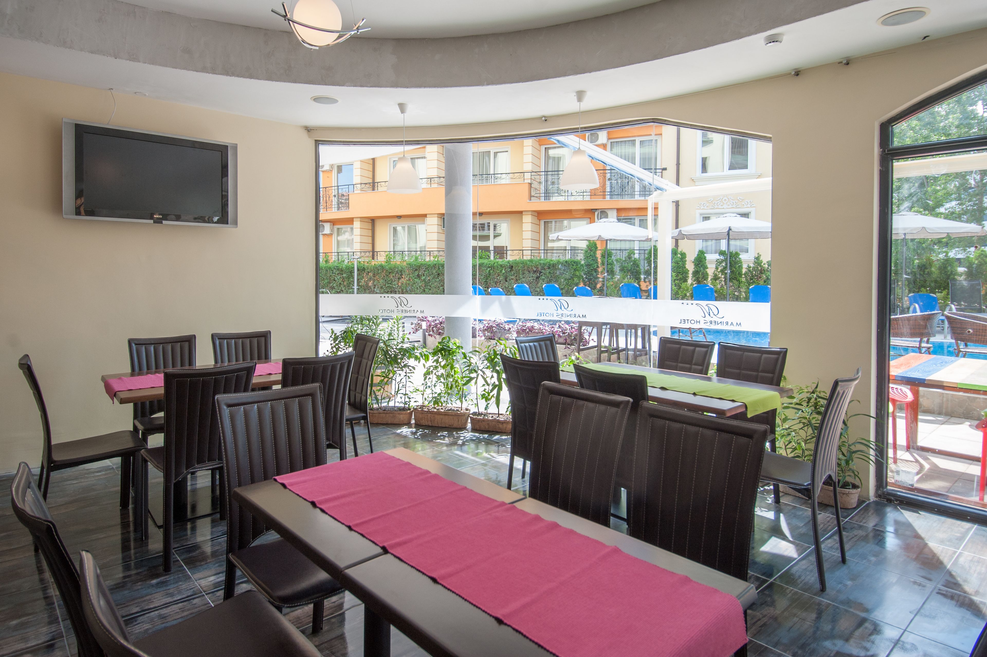 A restaurant with tables and chairs, a TV mounted on the wall, and a view of a building with balconies.