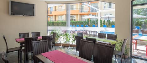 Un restaurante con mesas y sillas, un televisor montado en la pared y vistas a un edificio con balcones.