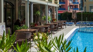 An outdoor seating area with wicker chairs and tables, a swimming pool, and a building with balconies in the background. - Mariner's Suites (Sunny Beach)