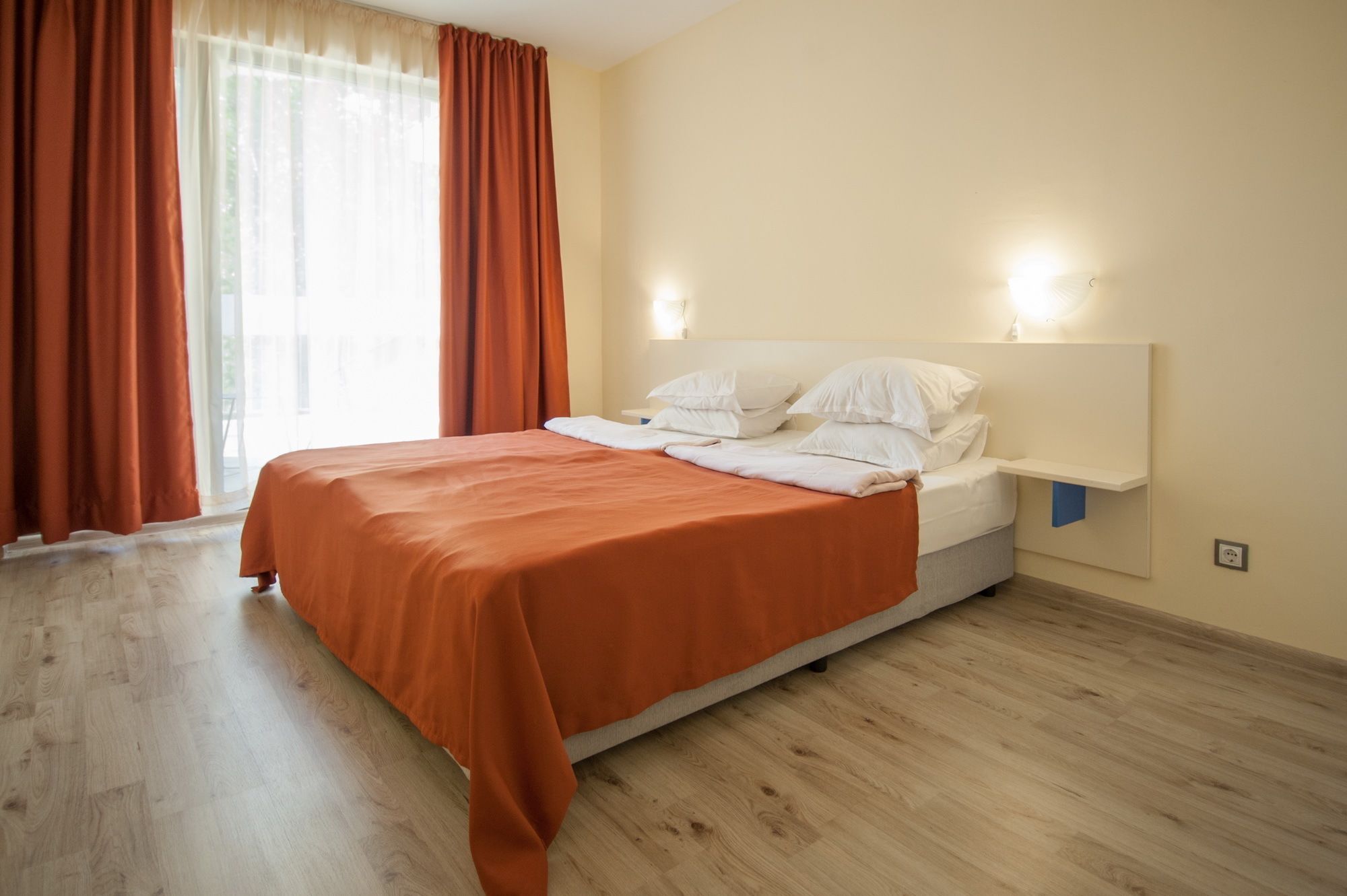 A neatly made bed with an orange bedspread, a white headboard, and two wall-mounted lights.