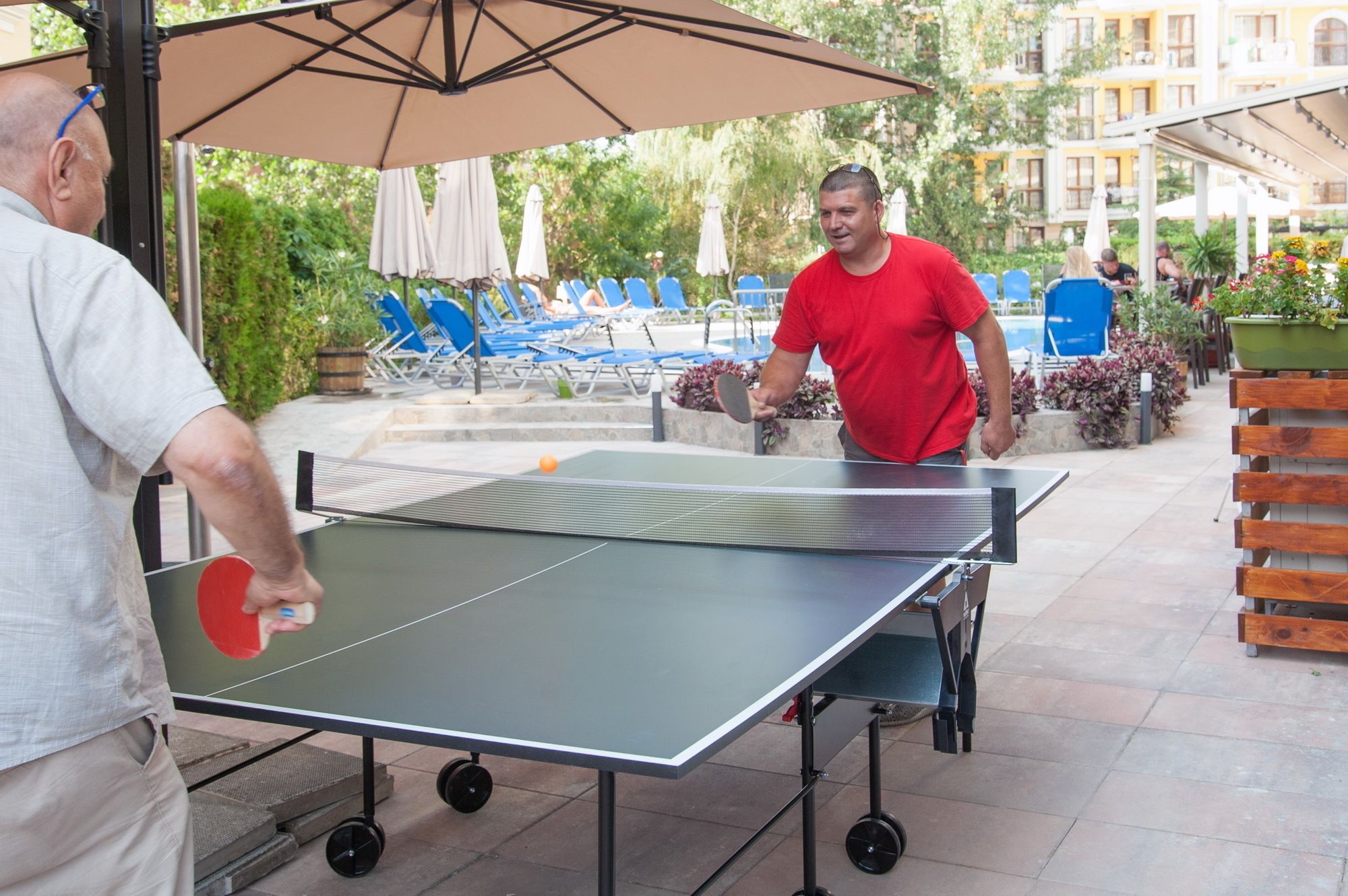 An outdoor ping pong table set up in a courtyard with lounge chairs and umbrellas in the background.