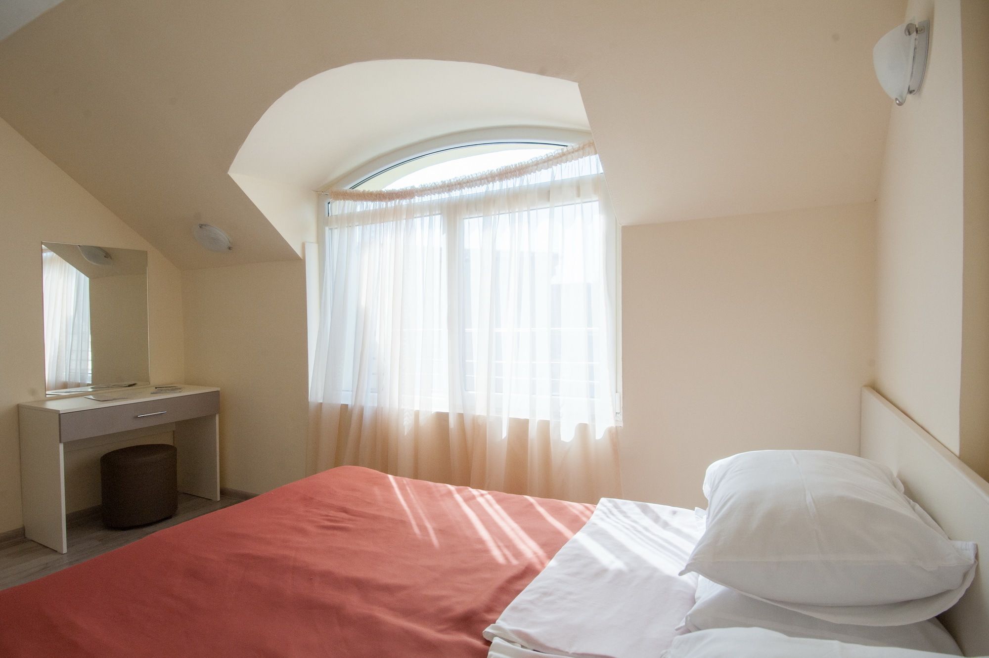 A bedroom with a bed, a bedside table, a mirror, a window with sheer curtains, and a sloped ceiling.
