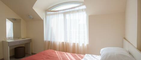 A bedroom with a bed, a bedside table, a mirror, a window with sheer curtains, and a sloped ceiling.