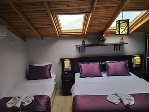 A room with two beds, each with purple bedding and pillows, a nightstand with a lamp, a shelf with decorative items, and a skylight above.