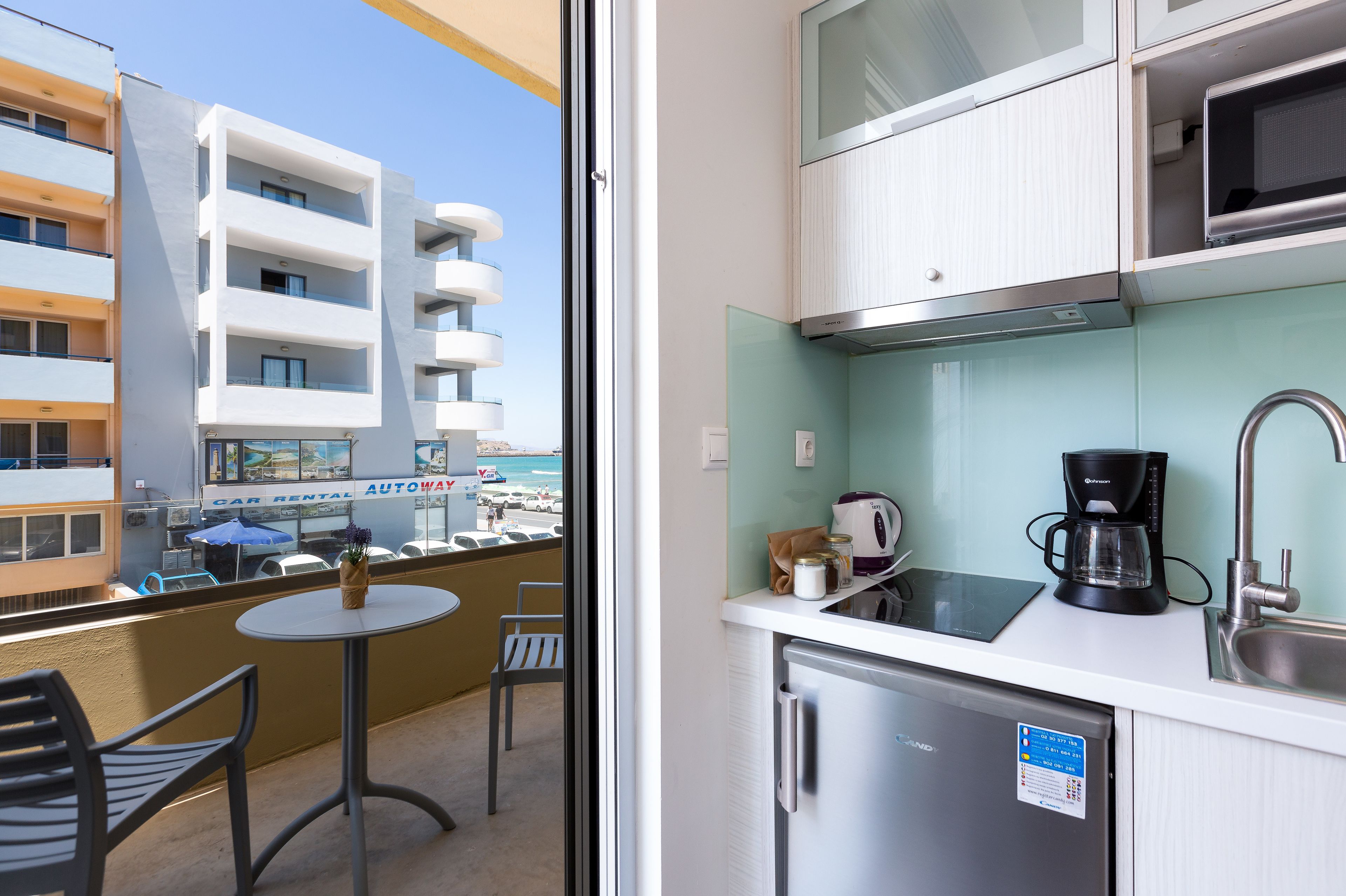 A balcony with a table and chairs, a view of a building with a car rental sign, and a kitchenette with a coffee maker and a refrigerator.