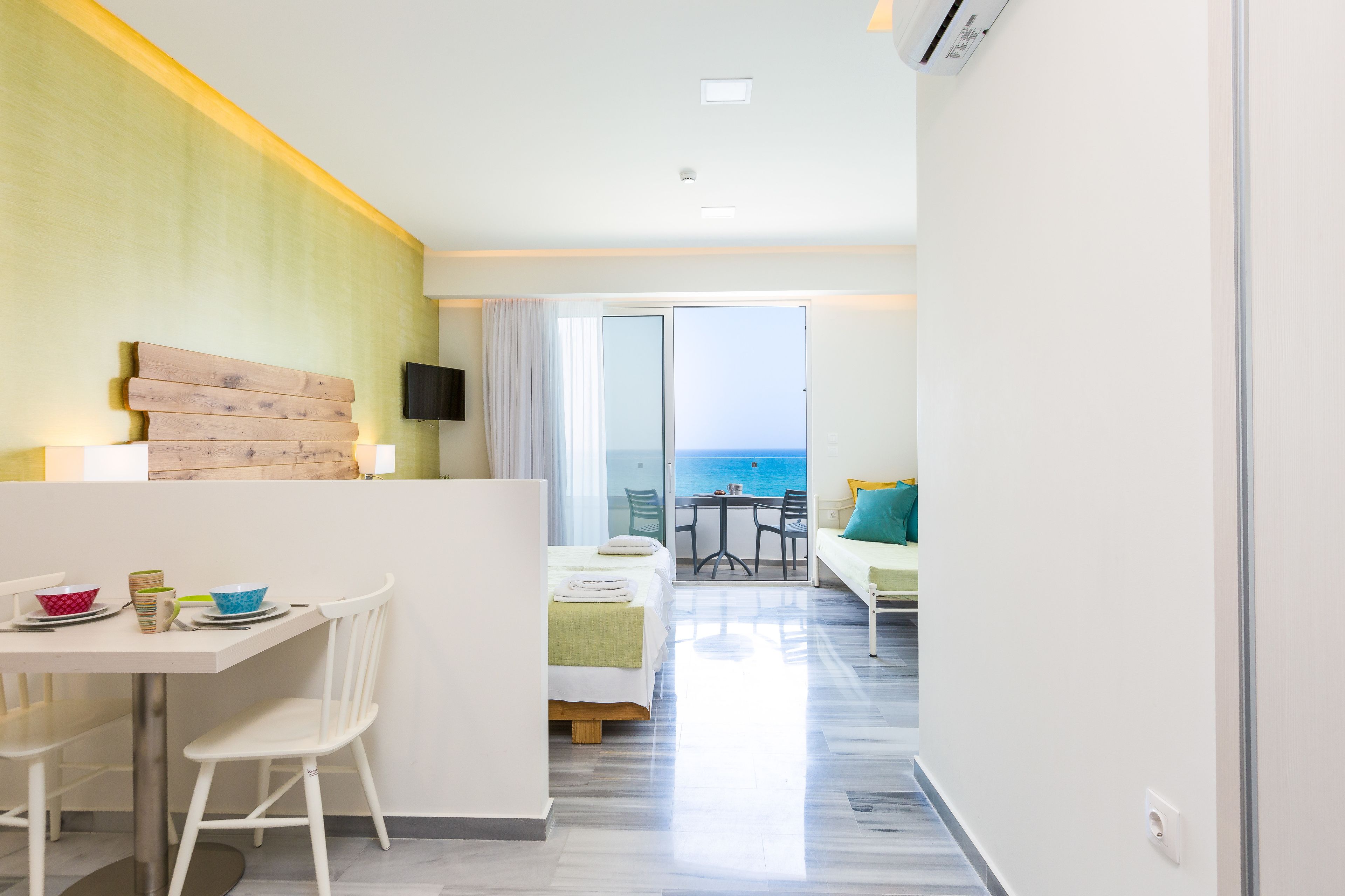 A hotel room with a bed, a dining area with a table and chairs, a TV mounted on the wall, and a view of the sea through a sliding door.