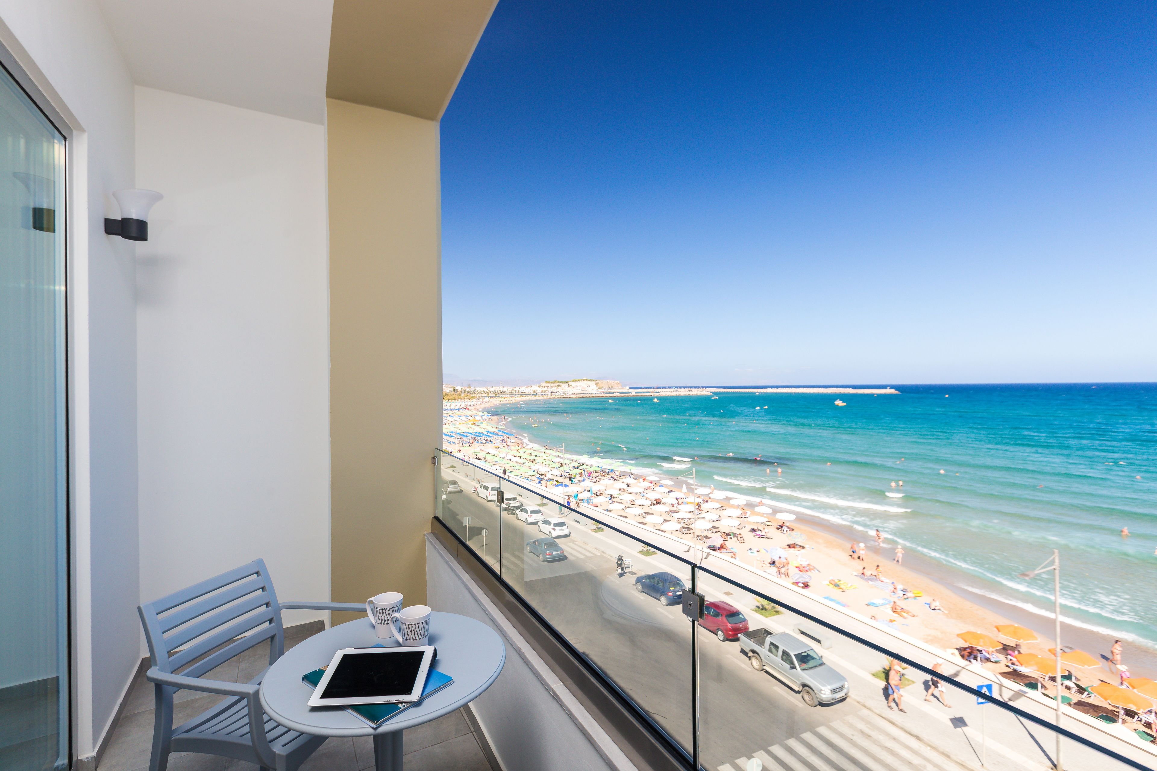 A balcony with a table, chair, and a tablet. The balcony overlooks a beach with umbrellas and people.
