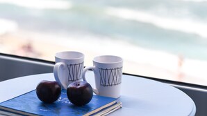A table with two white mugs with black designs, two dark-colored apples, and a blue book on a white table by a window overlooking the ocean. - Meltemi Coast Suites (Rethymno)