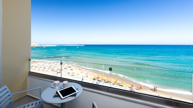 A balcony with a view of a beach, a table with a tablet and a mug, and a chair. - Meltemi Coast Suites (Rethymno)