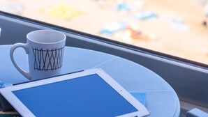 A tablet and a mug on a table by a window overlooking a beach. - Meltemi Coast Suites (Rethymno)