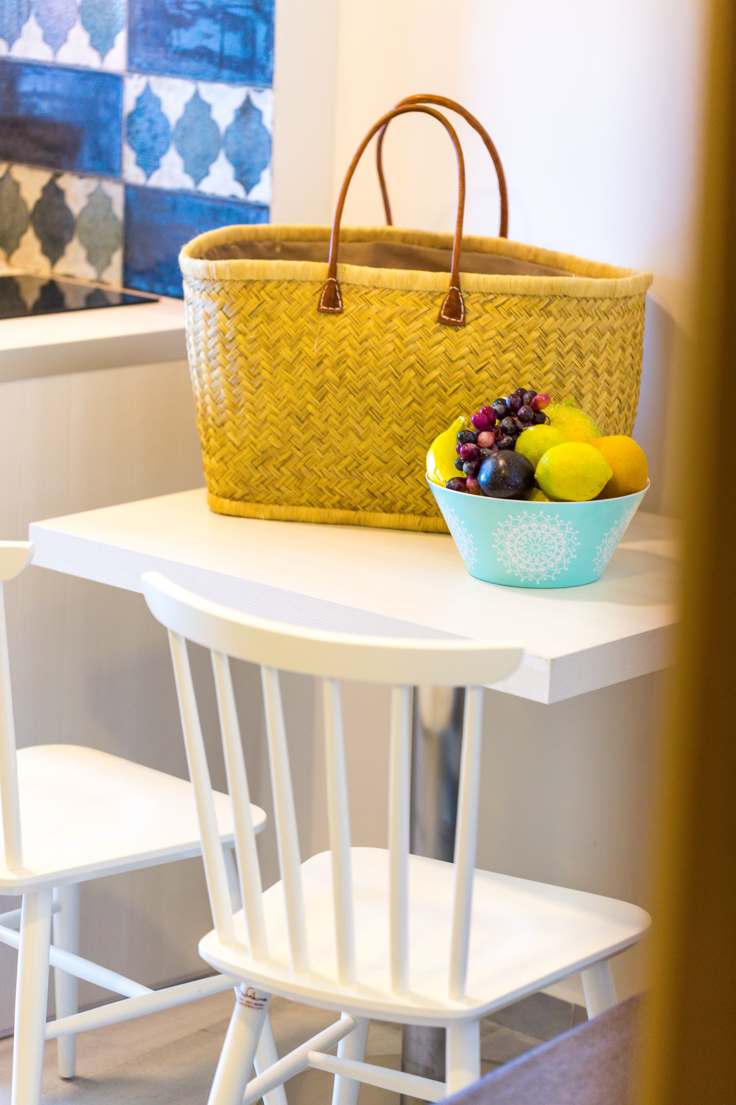 Una cesta amarilla tejida con asa sobre una mesa blanca. Junto a ella, un cuenco azul con frutas.