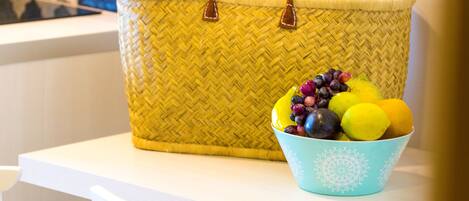 Una cesta amarilla tejida con asa sobre una mesa blanca. Junto a ella, un cuenco azul con frutas.