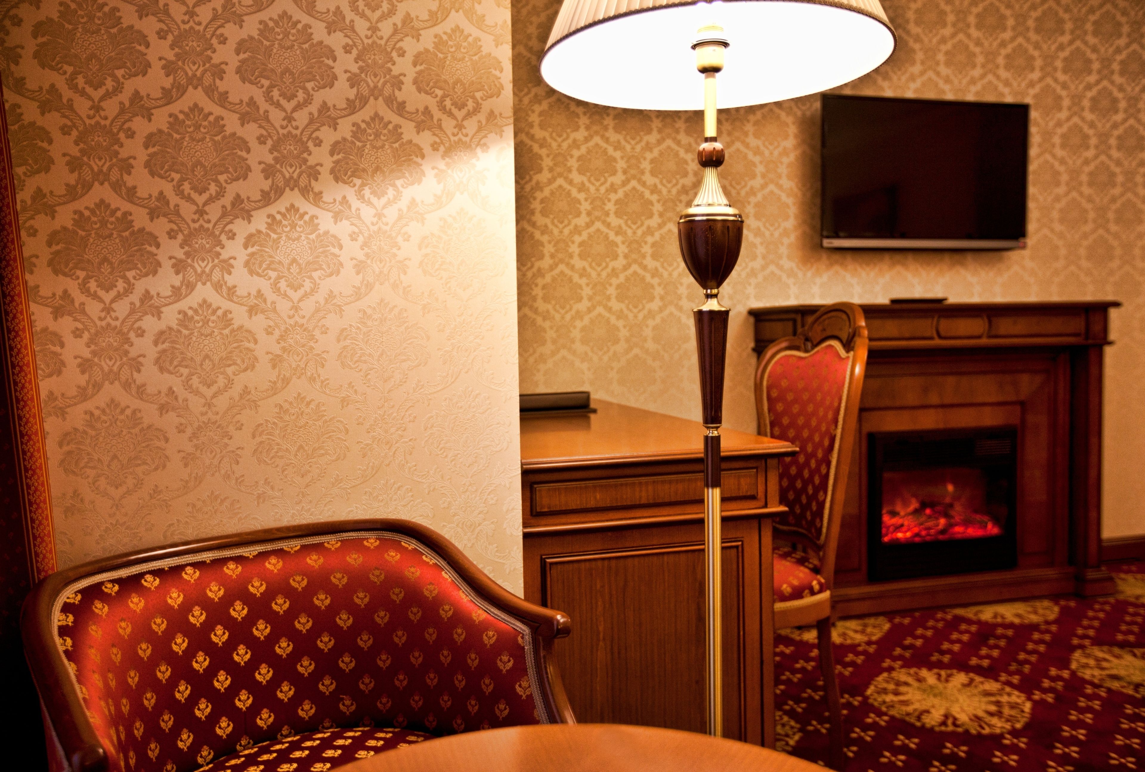 A room with a patterned wallpaper, a wooden chair, a lamp, a television, and a fireplace.