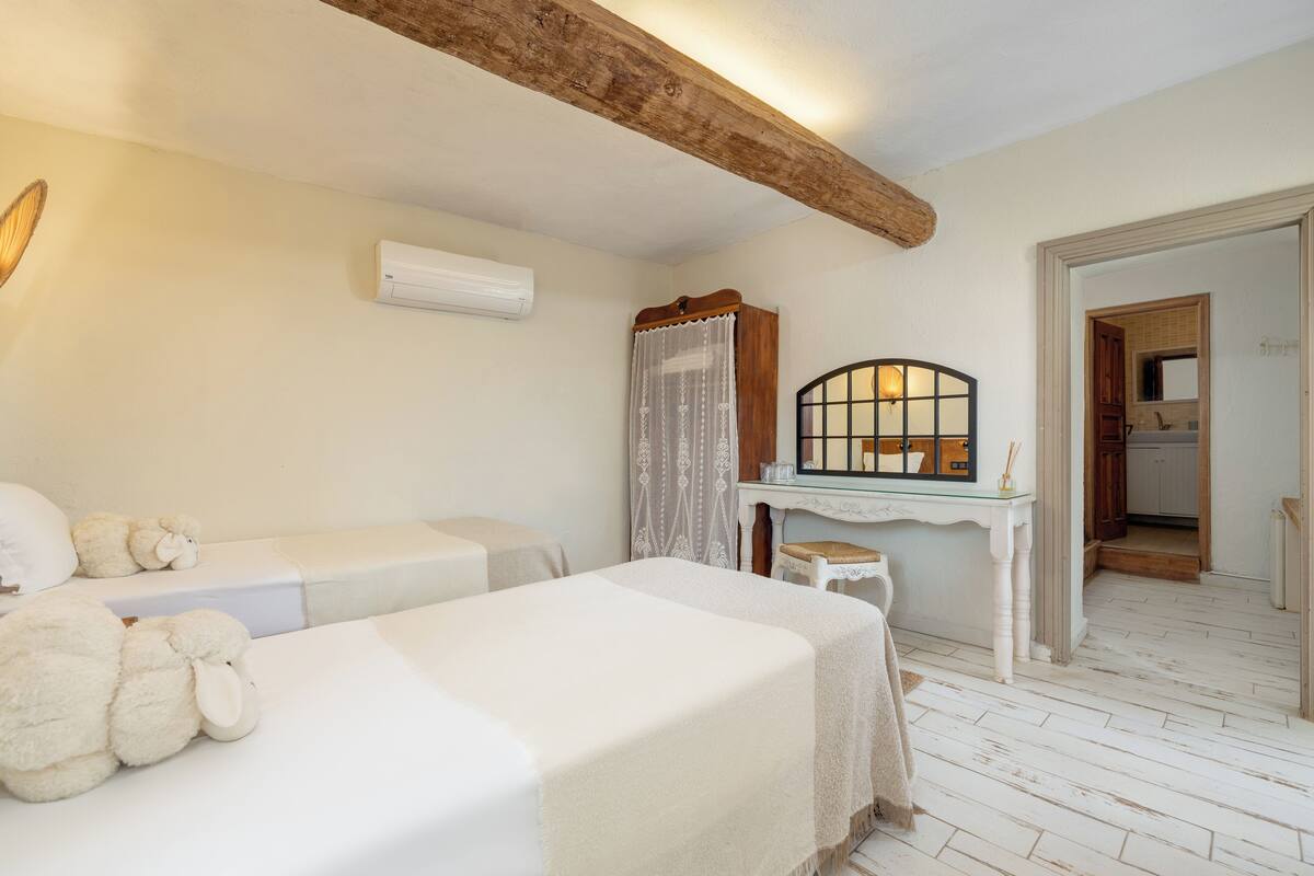 A bedroom with two beds, a wooden ceiling beam, a small table with a mirror, and a doorway leading to a bathroom.