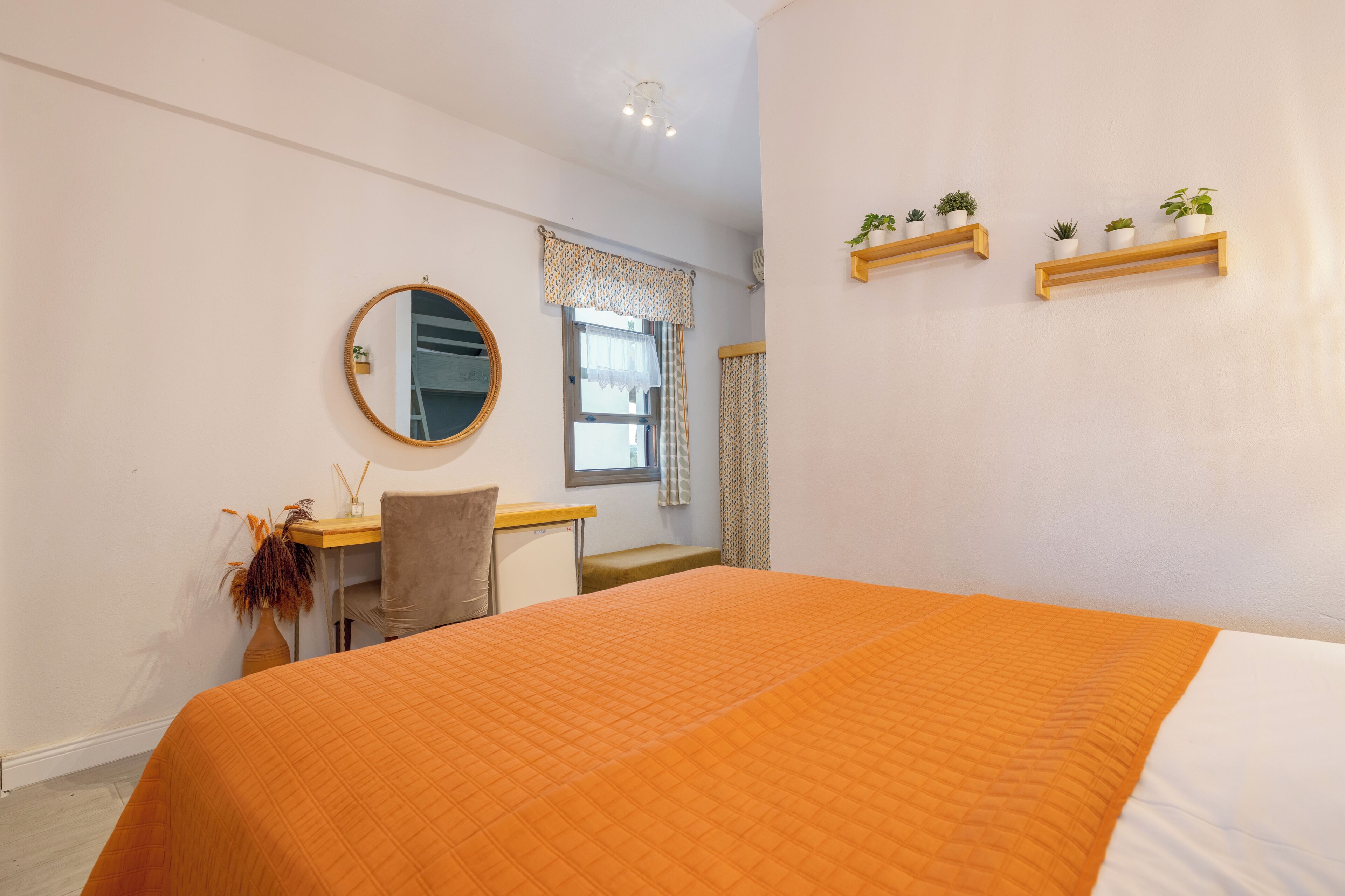 A bedroom with a large bed, a desk with a chair, a mirror, and two shelves with plants.