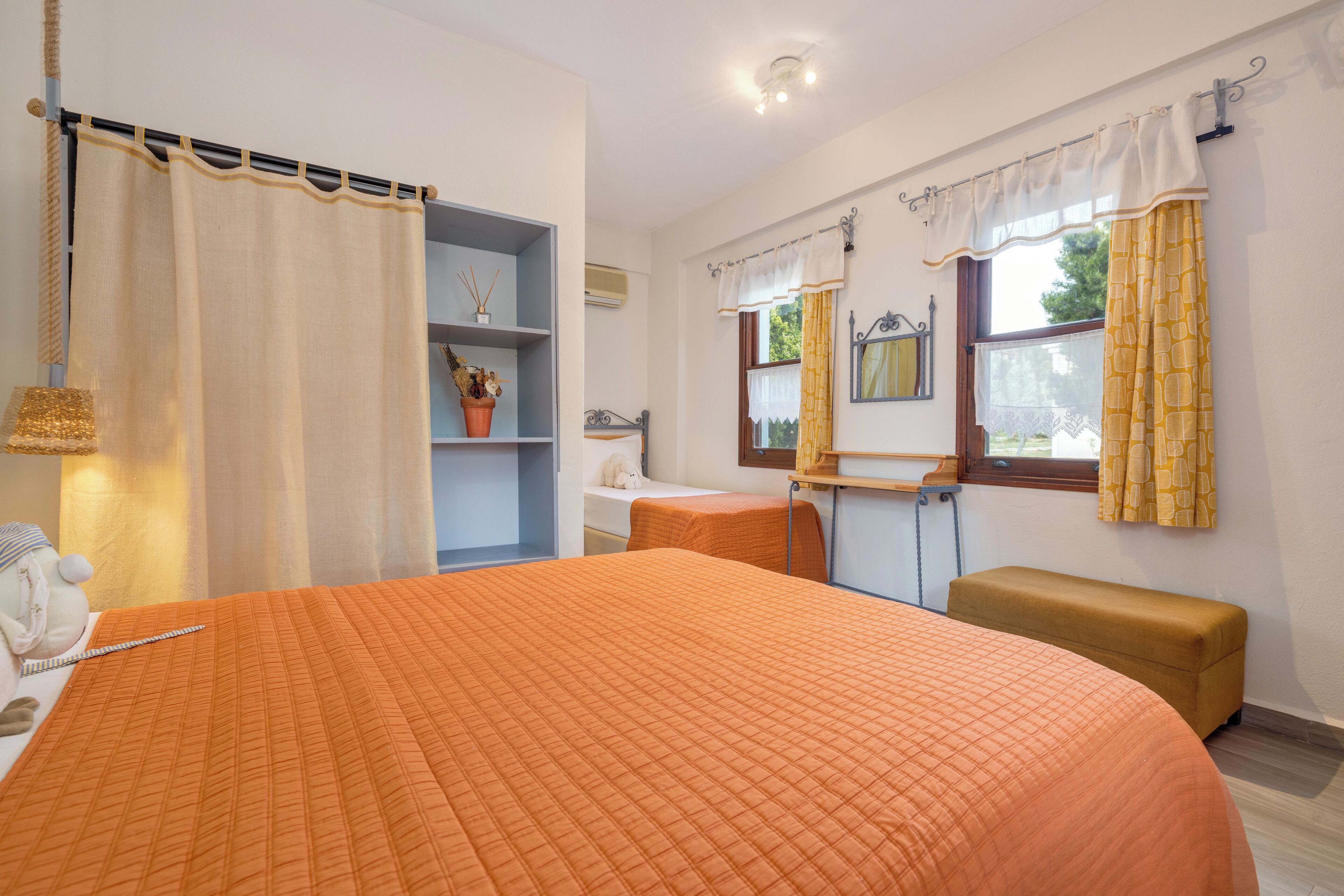 A bedroom with a large bed, a bedside table, a bench, a shelf with decorative items, and a window with curtains.