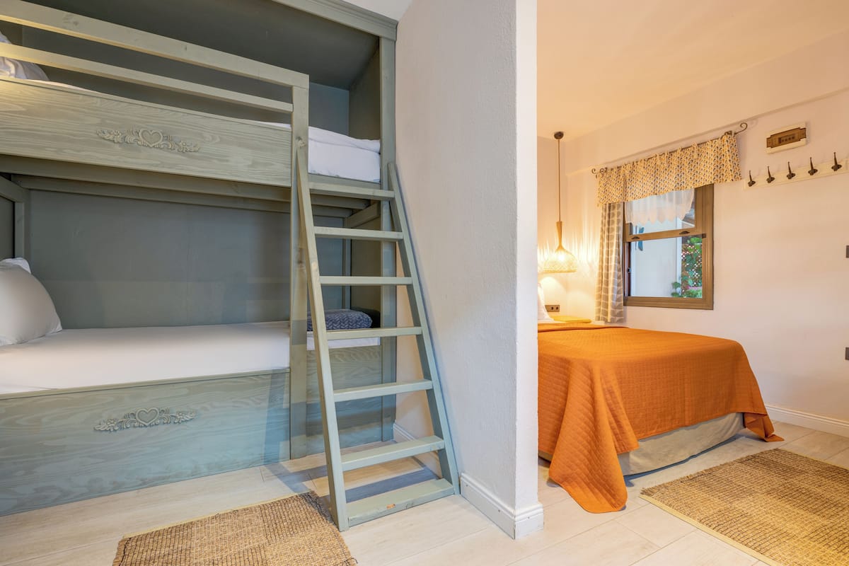 A bedroom with a bunk bed, a ladder, a bedside table with a lamp, and a window with a curtain.