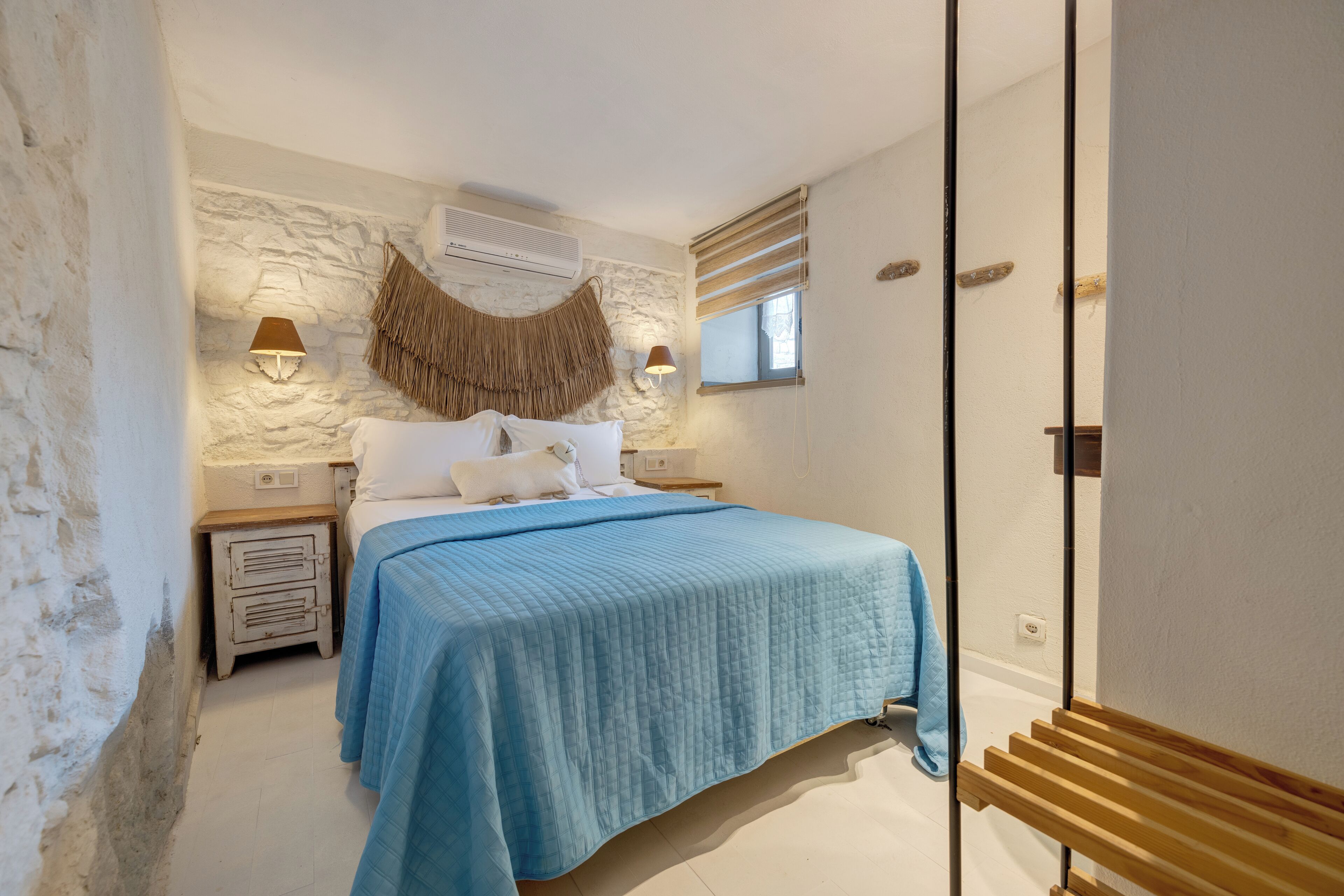 A bedroom with a stone wall, a bed with a blue bedspread, two bedside lamps, and a window with blinds.