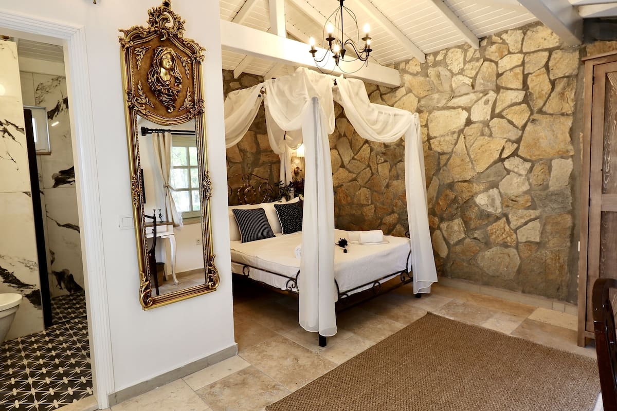 A bedroom with a stone wall, a canopy bed, a large ornate mirror, and a chandelier.