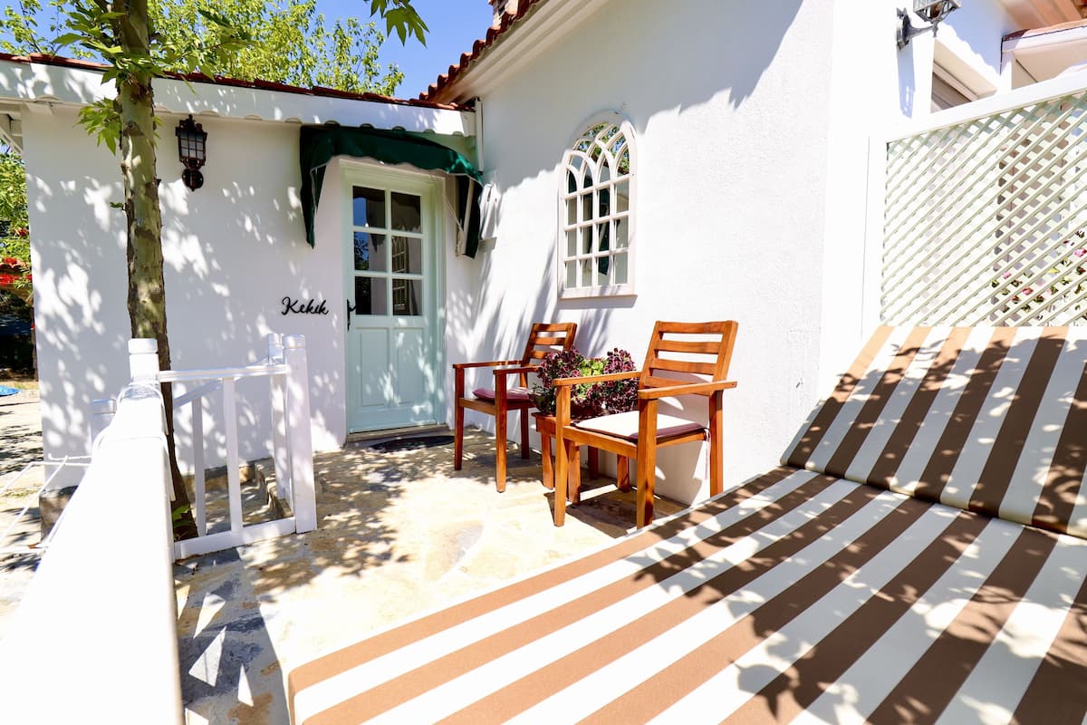 A white building with a green awning, a wooden bench, and two wooden chairs with cushions.