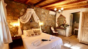 Un dormitorio con pared de piedra, cama con dosel, mesita de noche con lámpara, una silla y baño visible a través de una puerta abierta.
