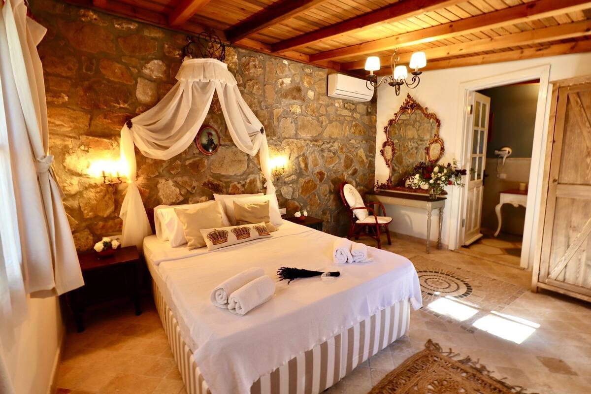 A bedroom with a stone wall, a canopy bed, a bedside table with a lamp, a chair, and a bathroom visible through an open door.