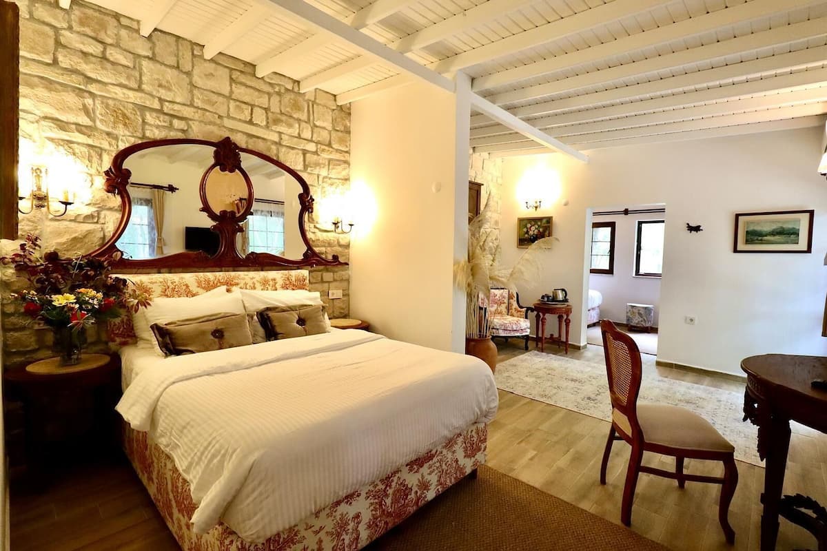 A bedroom with a stone wall, a large bed with floral bedding, a wooden chair, and a small table.
