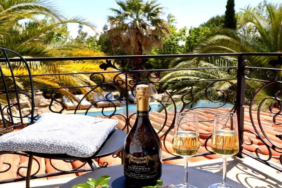 A balcony with a bottle of wine and two glasses on a table, overlooking a pool and palm trees.