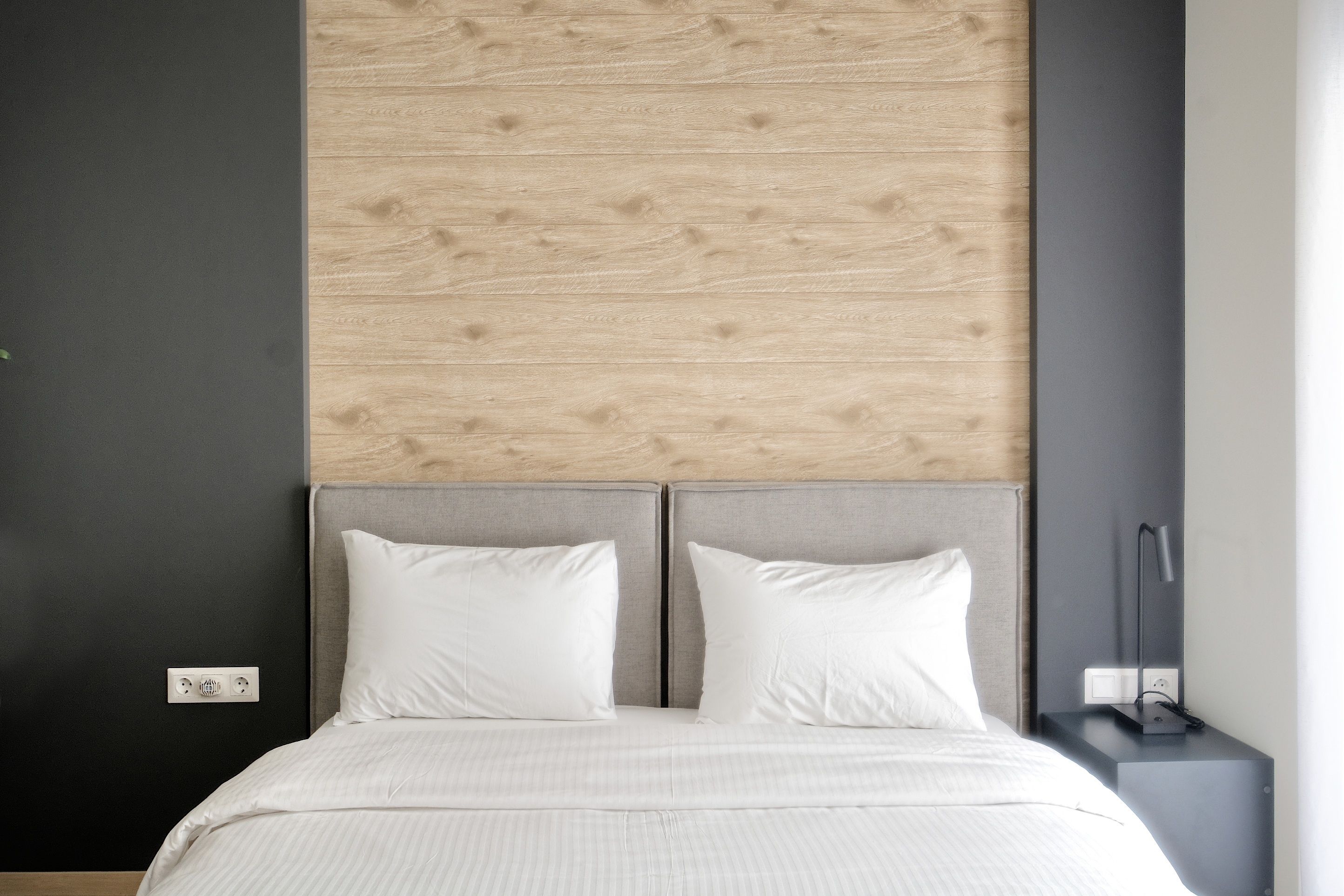 Une chambre à coucher moderne avec une tête de lit en bois, deux oreillers blancs, un lit recouvert d’une couverture à rayures et une table de chevet avec une lampe.