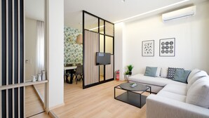 Una sala de estar moderna con un sofá, una mesa de centro y un televisor montado en la pared. Se observa un área de comedor con una mesa y sillas, separada por una partición.