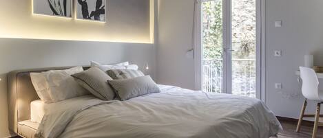 A bedroom with a bed, pillows, a desk, and a window with a view of greenery.