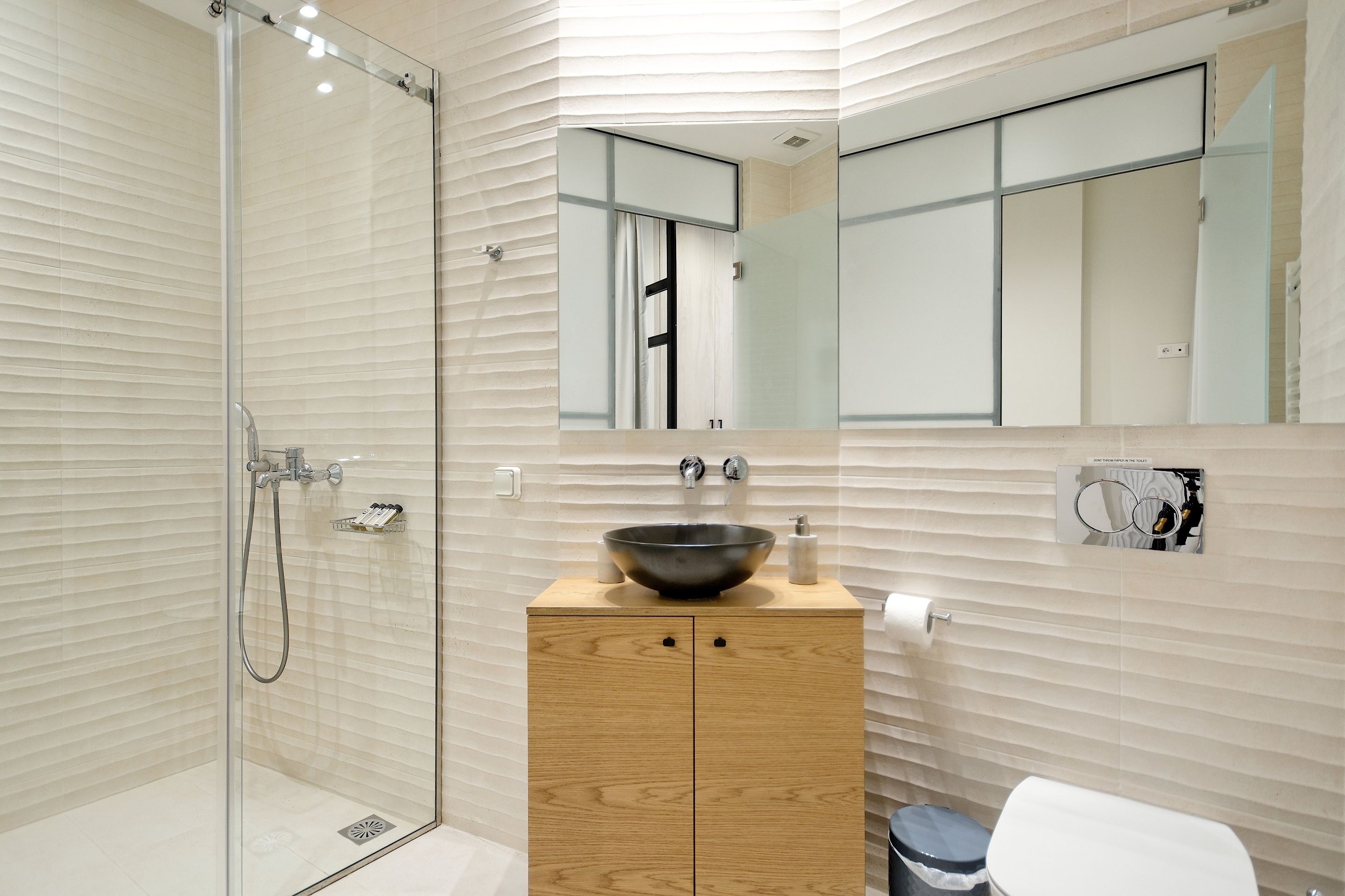 A modern bathroom with a glass-enclosed shower, a wooden vanity with a black vessel sink, and a large mirror above the sink.