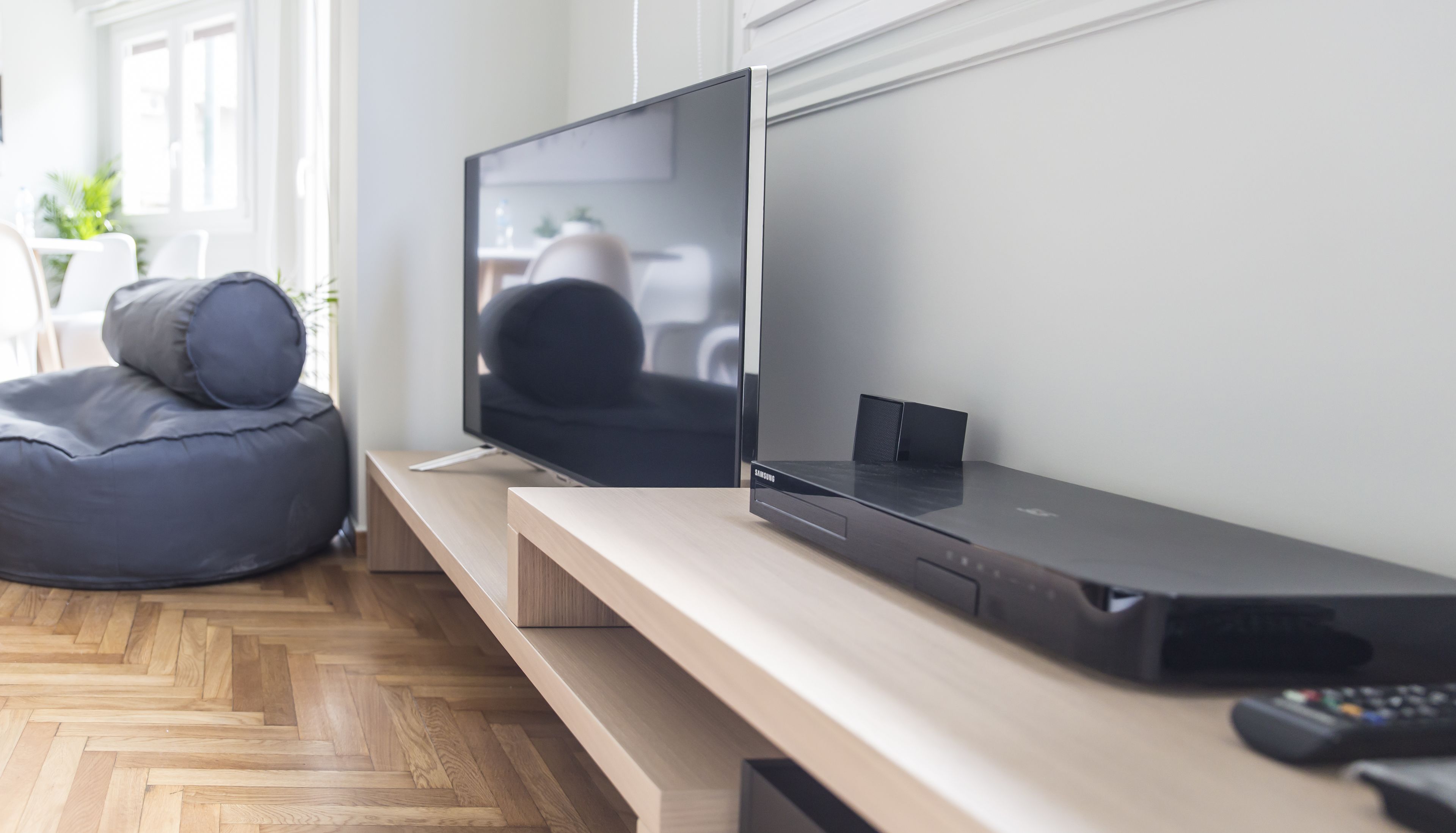 A modern living room with a flat-screen TV, a soundbar, and a gaming console on a wooden TV stand. There is a bean bag chair and a small table with a plant.