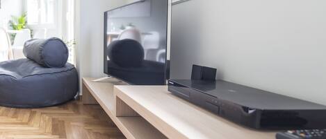 A modern living room with a flat-screen TV, a soundbar, and a gaming console on a wooden TV stand. There is a bean bag chair and a small table with a plant.