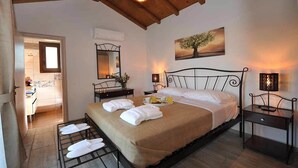 A bedroom with a large bed, a bedside table, a nightstand, a hanging light fixture, a painting, and a bathroom visible through an open door. - OLIVIA VILLAS (Ikaria)