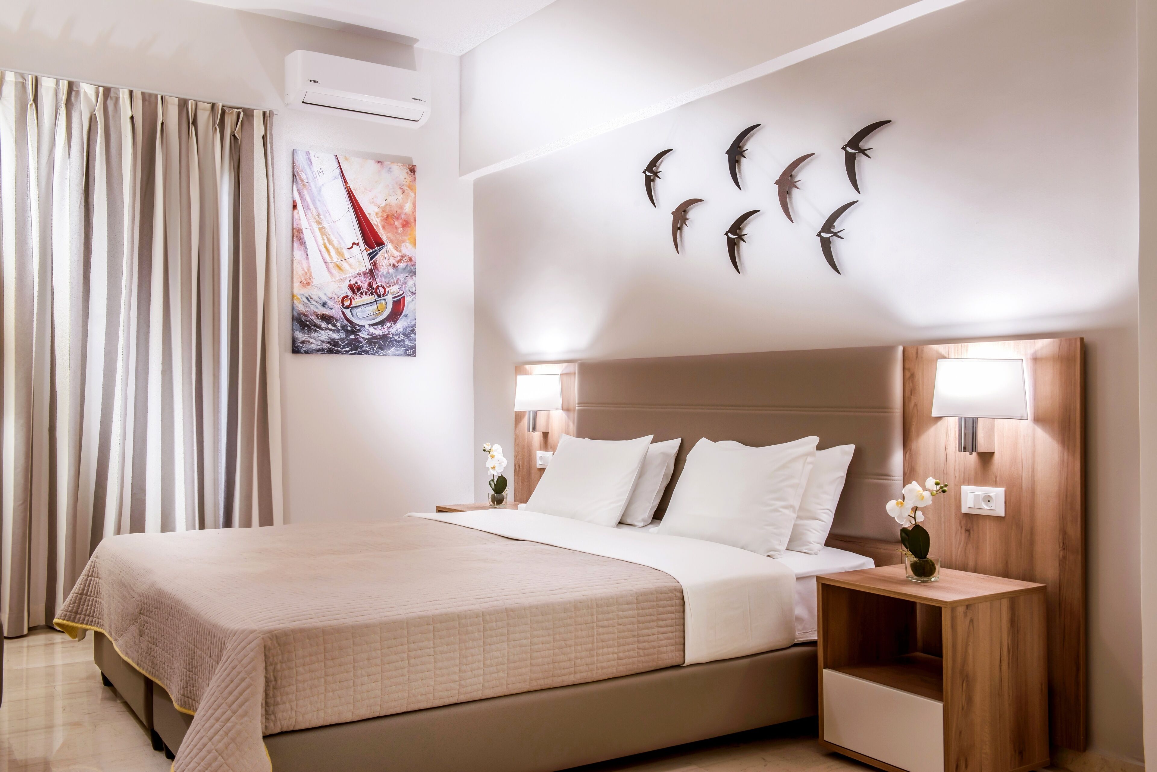 A hotel room with a bed, curtains, a painting, wall-mounted bird sculptures, and a nightstand with a lamp.