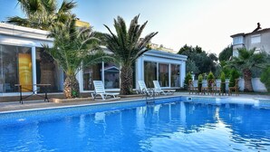 A swimming pool with lounge chairs, palm trees, and a building with large windows. - Uzunhan Hotel (Ayvalik)