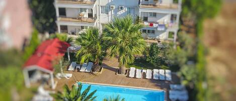 A two-story building with a swimming pool, sun loungers, and palm trees.