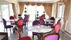 A dining area with round tables and chairs, large windows with curtains, and a view of the outdoors. - Uzunhan Hotel (Ayvalik)