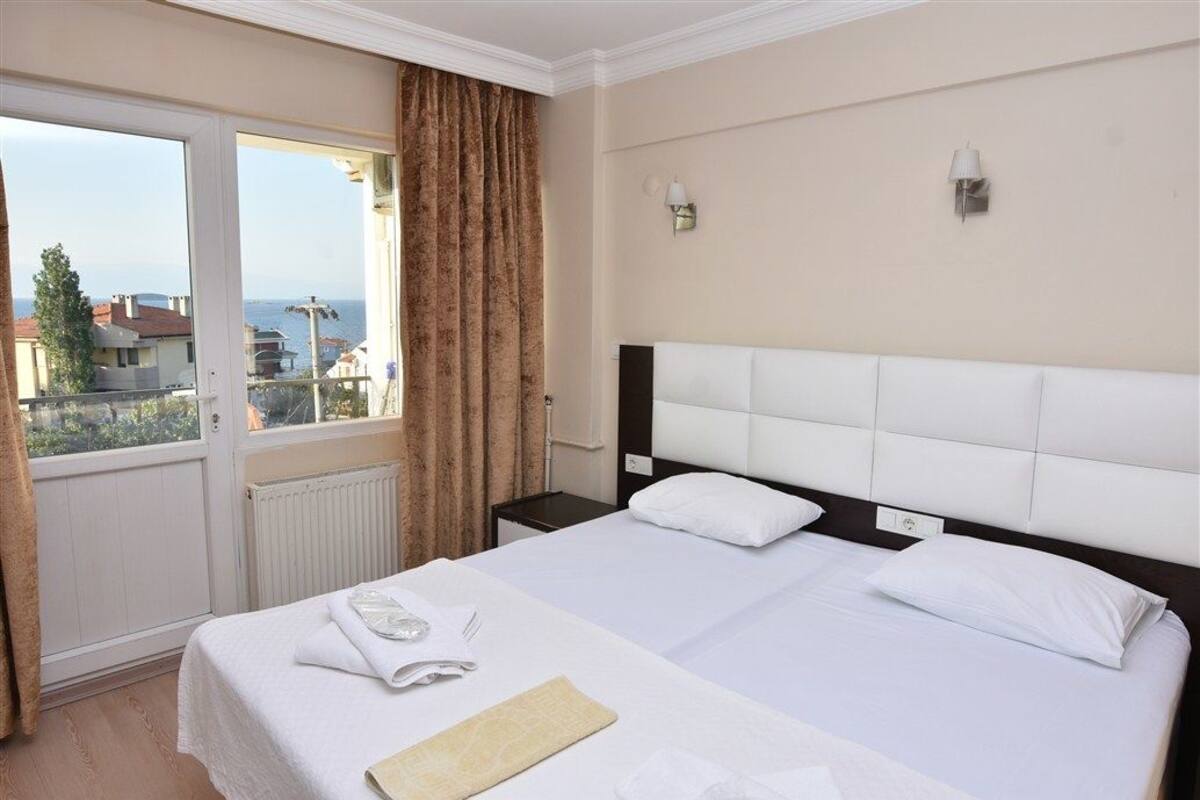 Une chambre d’hôtel avec un grand lit, une fenêtre avec des rideaux, une vue sur la mer et une tête de lit blanche.
